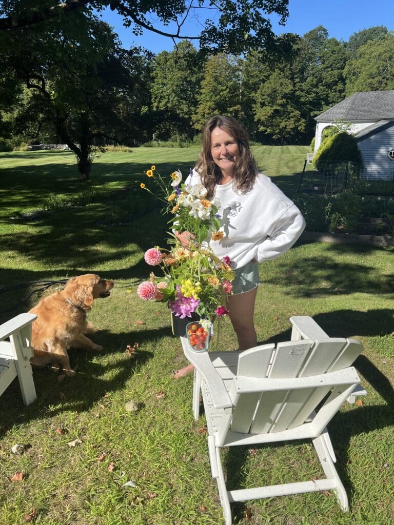 Woman with flowers beside dog and chairs.
